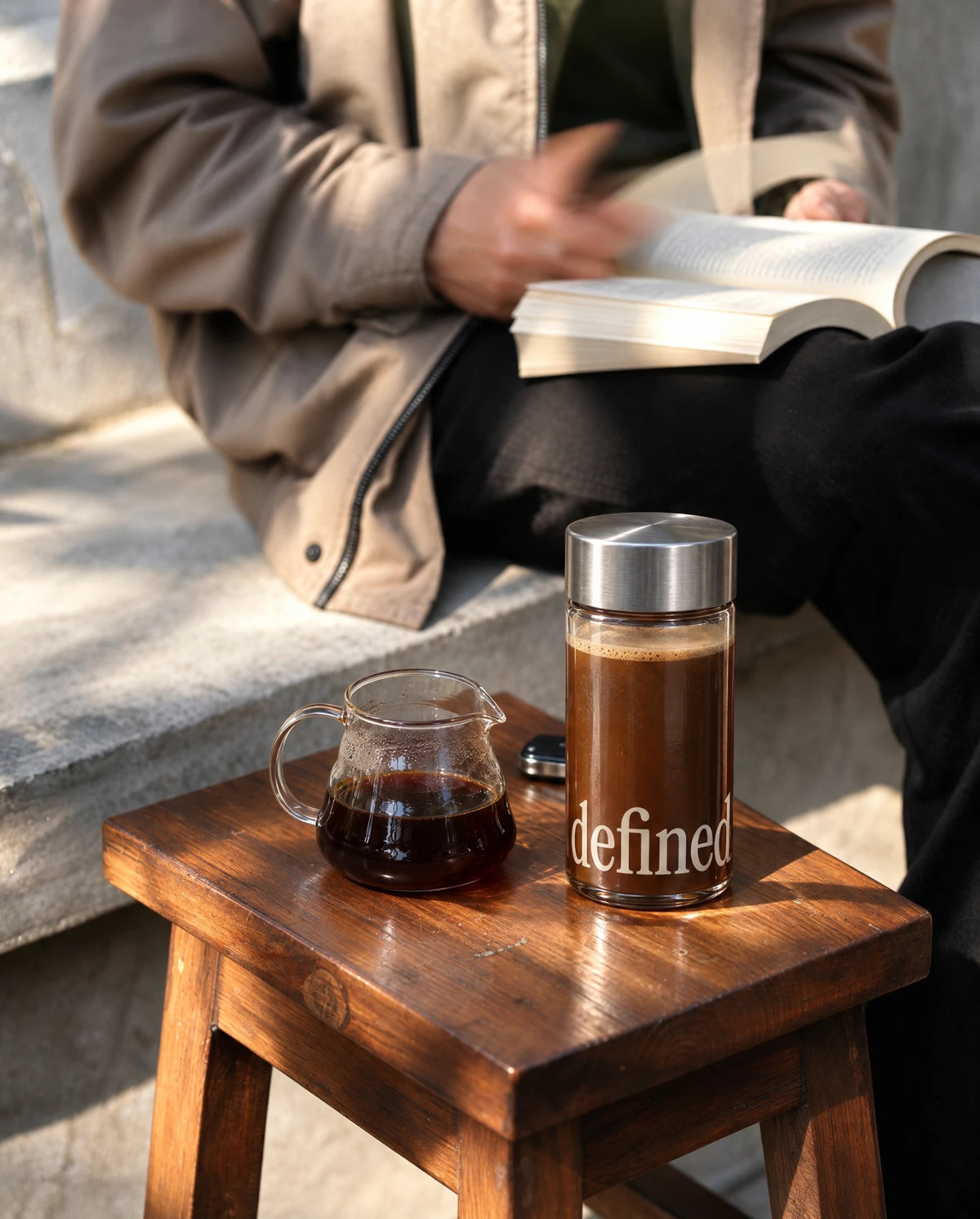 Bottle of defined black coffee and a glass carafe on a wooden stool beside someone reading