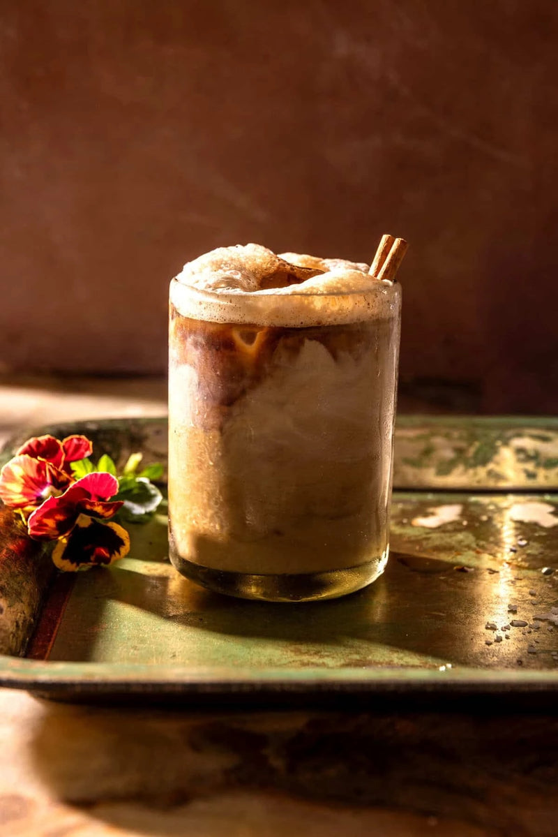 Foamy iced latte with a cinnamon stick on a green tray beside pansies